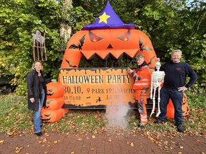 Mitarbeiter der Stadt und der stellvertretende Ortsbrandmeister stehen vor dem Werbebanner für die Halloween-Party