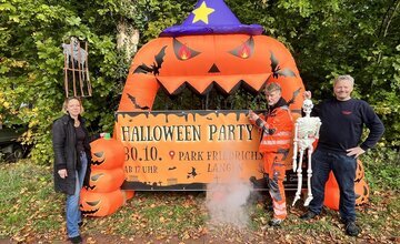 Mitarbeiter der Stadt und der stellvertretende Ortsbrandmeister stehen vor dem Werbebanner für die Halloween-Party